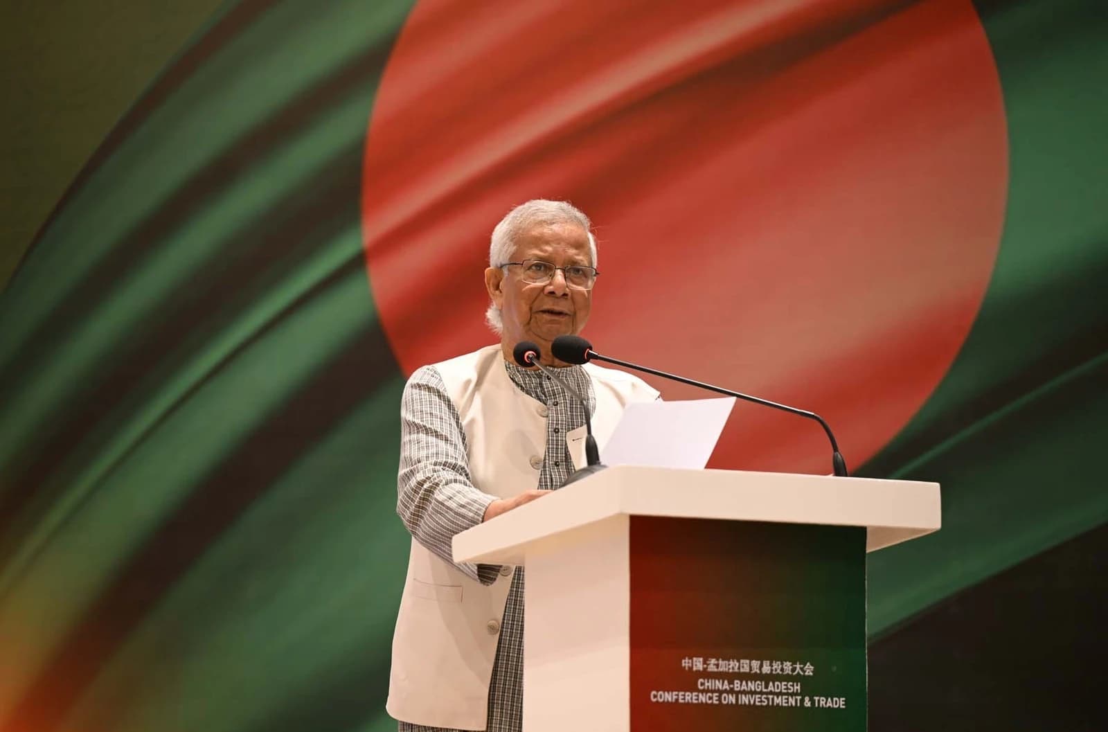 Muhammad Yunus speaking at a podium with microphones at an investment conference.
