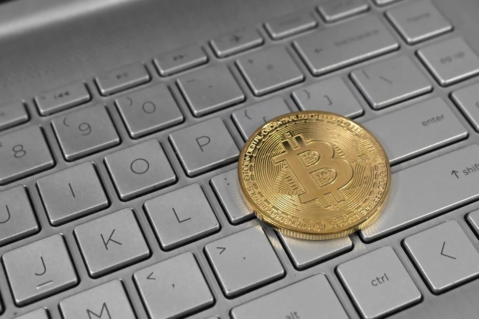 A physical golden Bitcoin cryptocurrency coin resting on a silver laptop keyboard.