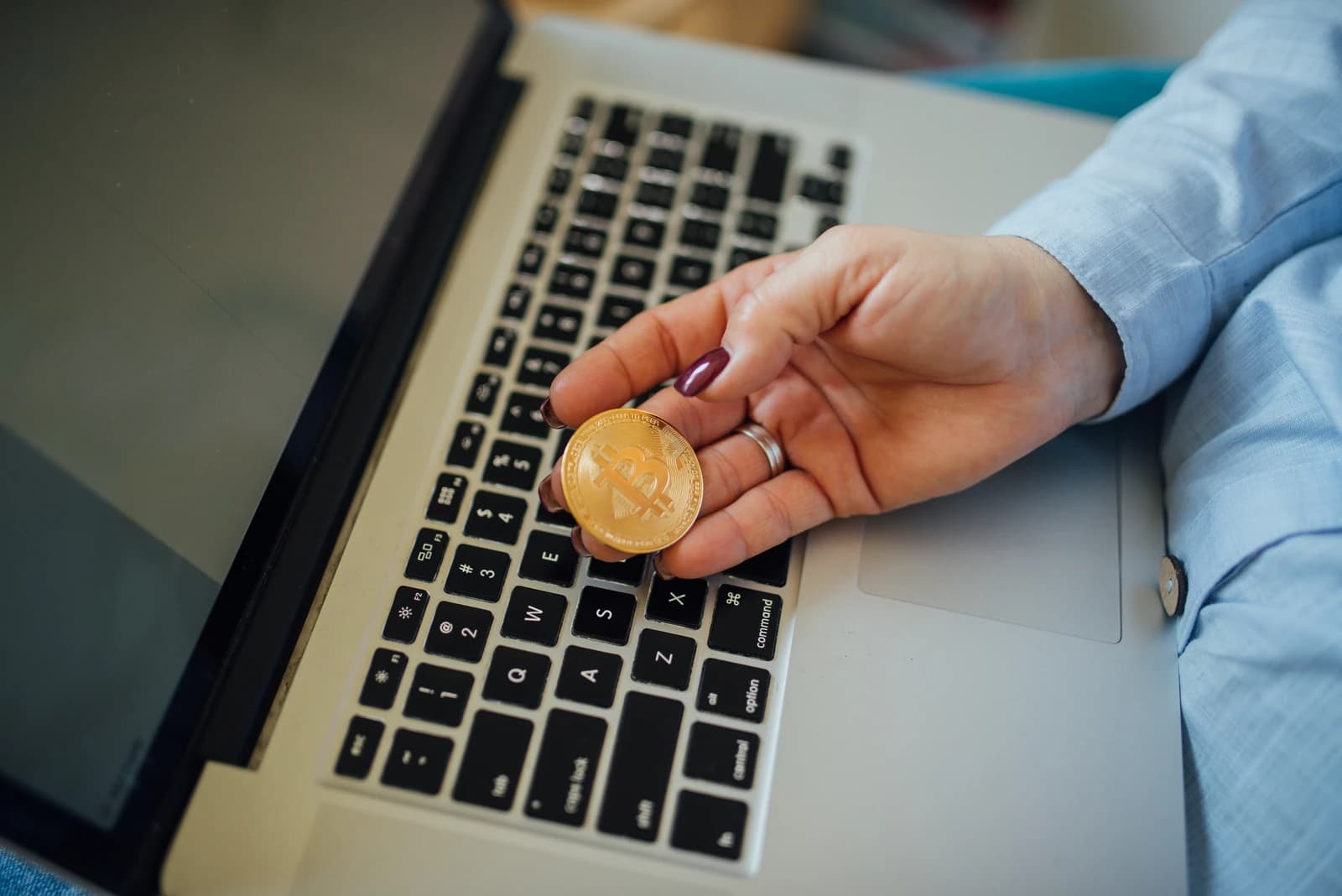 Woman's hand holding a physical Bitcoin coin above a laptop keyboard.