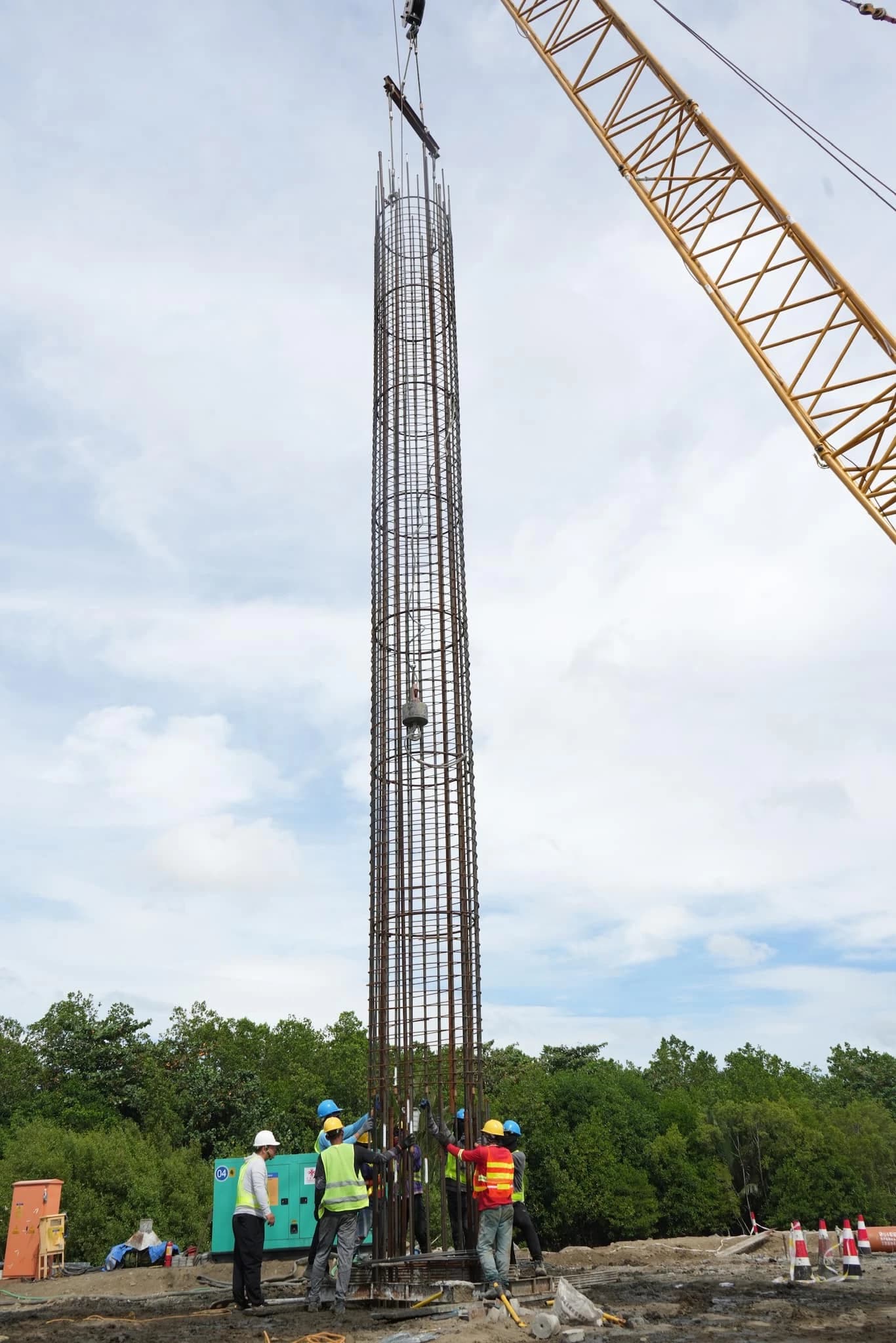 Construction workers guide large iron piling being lowered by a crane at a bridge construction site.