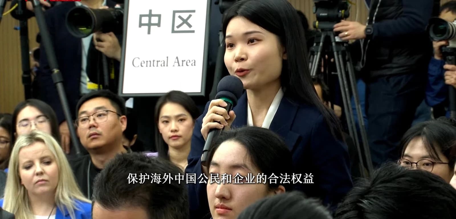 China News Service journalist speaking into a microphone at an official event.