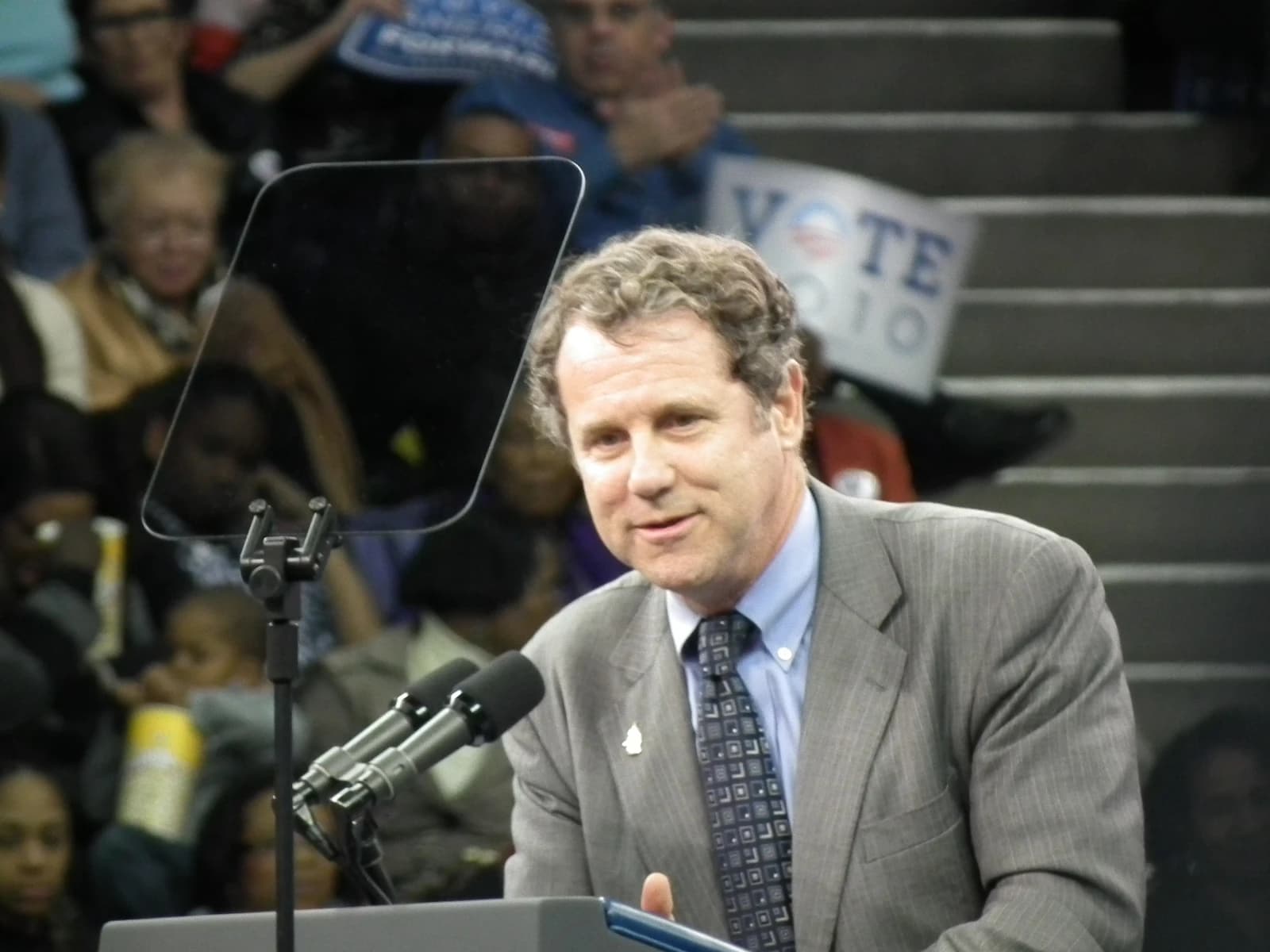 Senator Sherrod Brown speaking at a podium with microphones in Cleveland, Ohio.