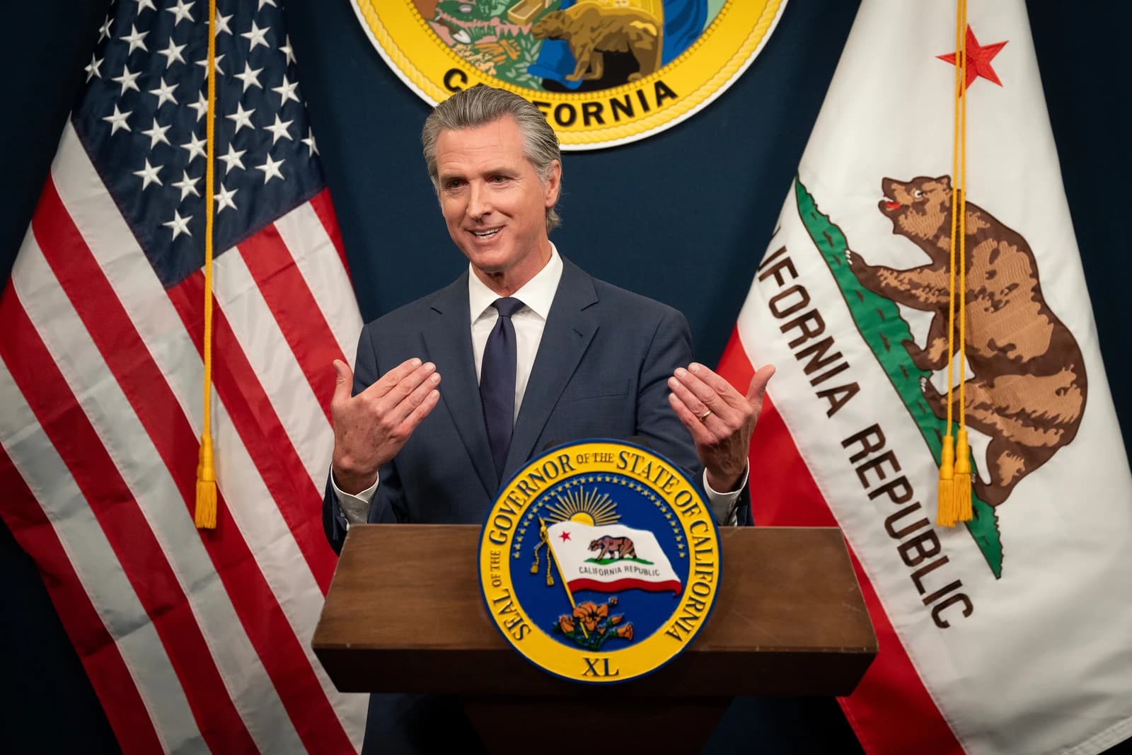 Governor Gavin Newsom speaking at a press conference in front of flags.