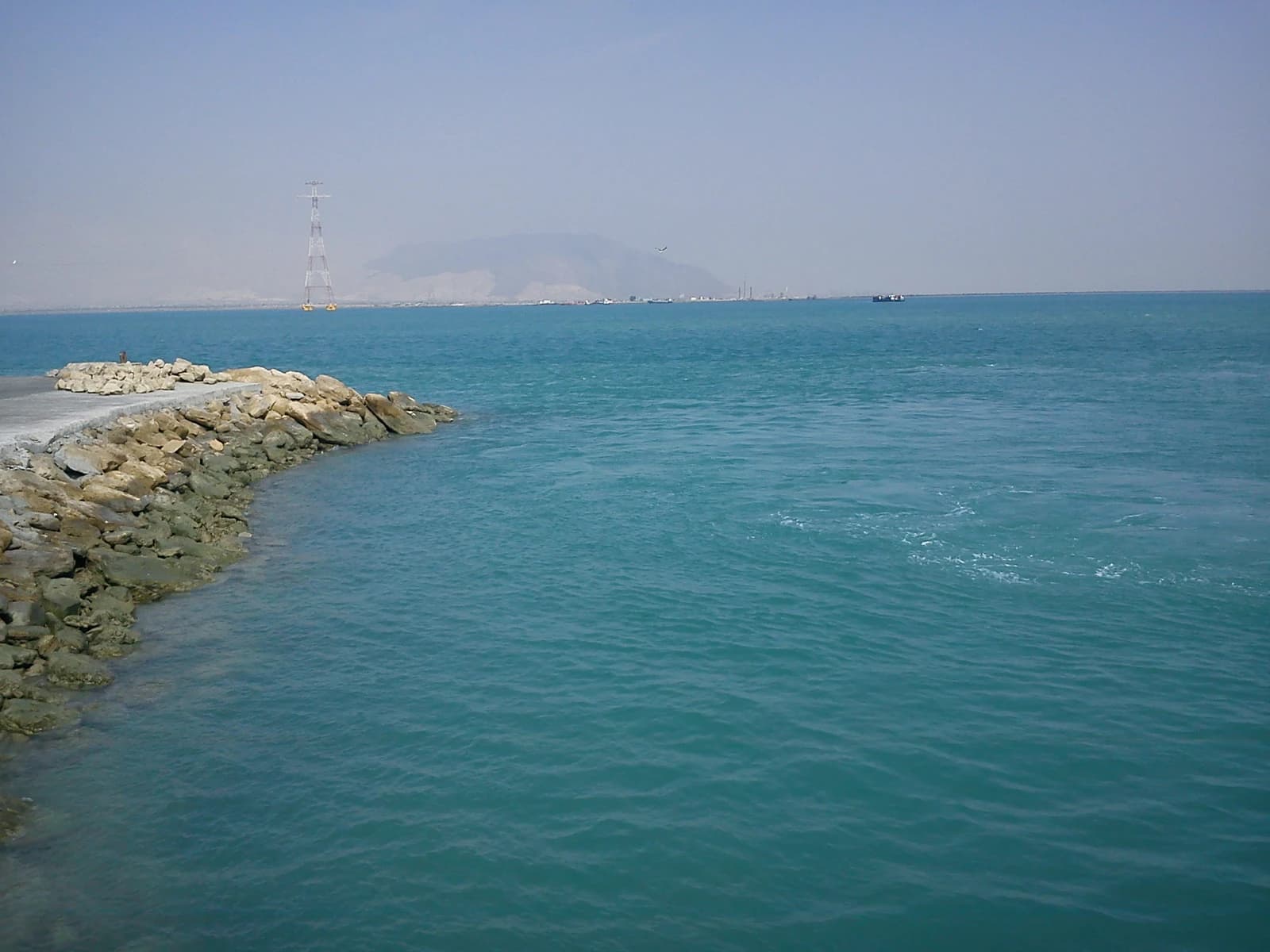 Transmission tower standing over water near a rocky coastline, with distant mountains.