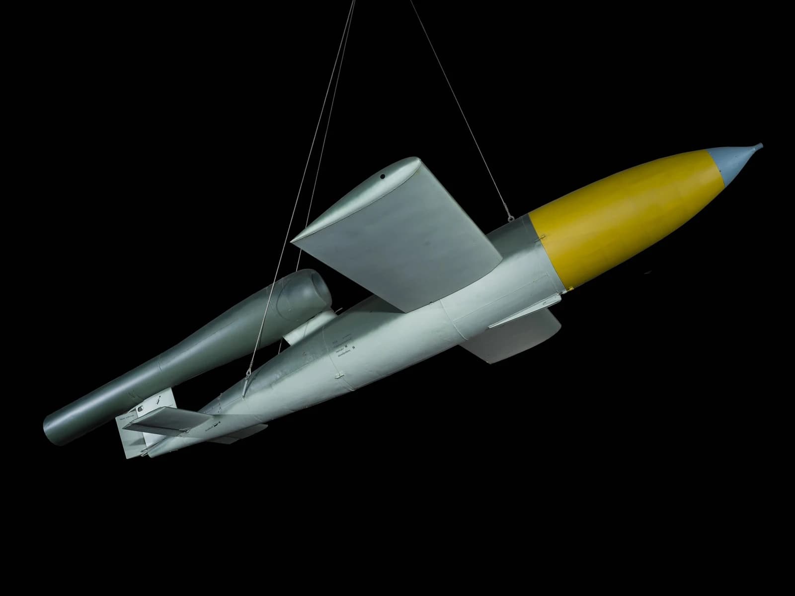 V-1 cruise missile with yellow nose cone suspended from wires against a black background.