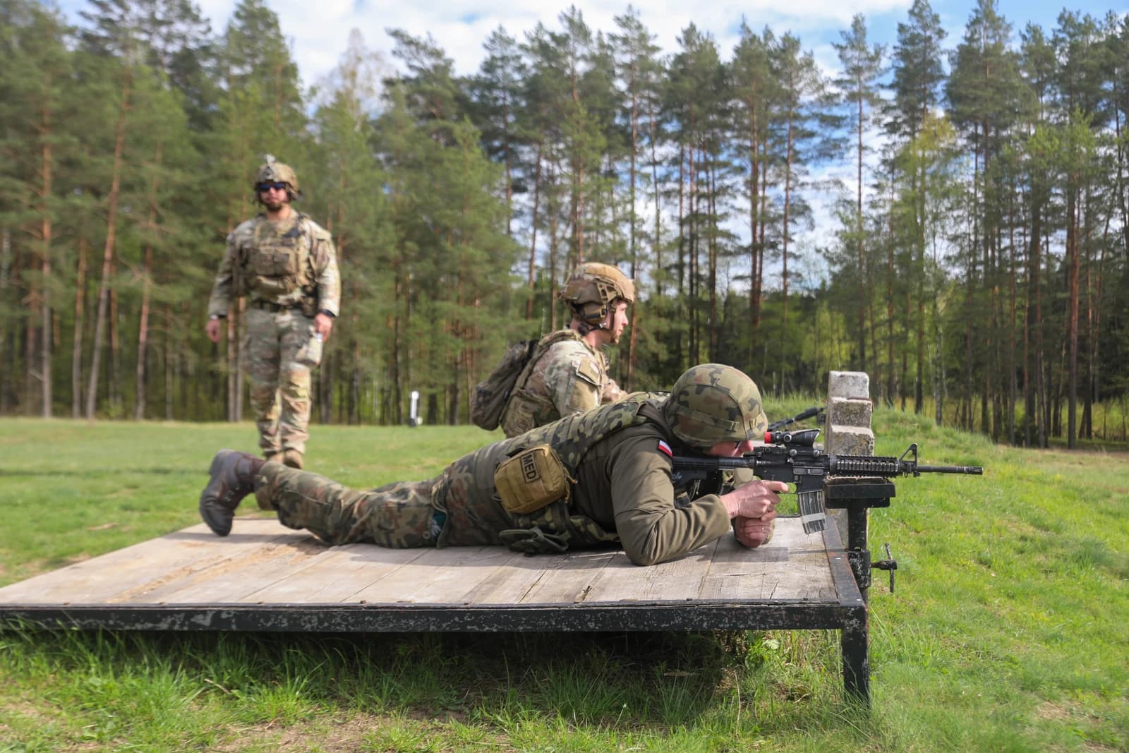 Soldier in camouflage uniform aiming a rifle from a prone position on a wooden platform.