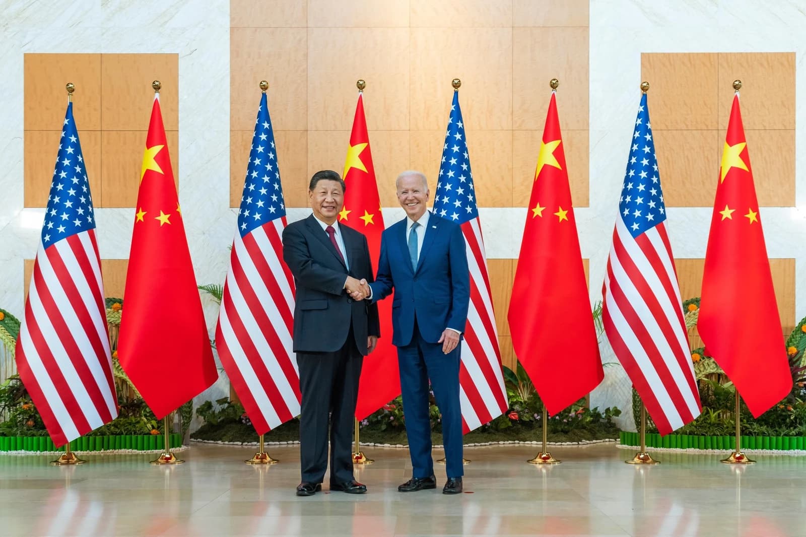 President Xi Jinping and President Joe Biden shaking hands in front of US and Chinese flags.