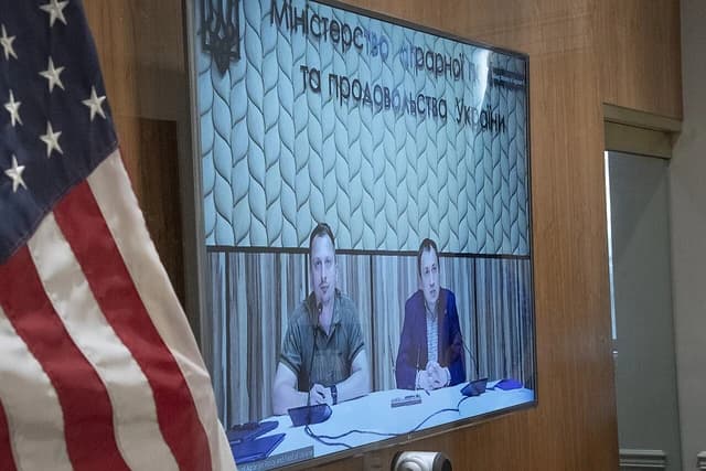 Ukrainian officials speaking during a video conference, with US flag in foreground.