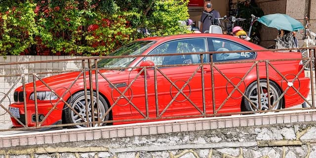 Red BMW 3 Series Coupe (E46) parked behind a rusted metal fence.