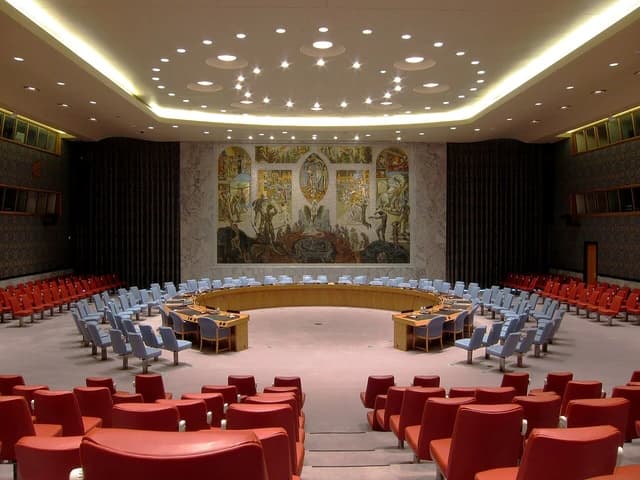 The United Nations Security Council chamber with empty chairs and a large mural on the wall.