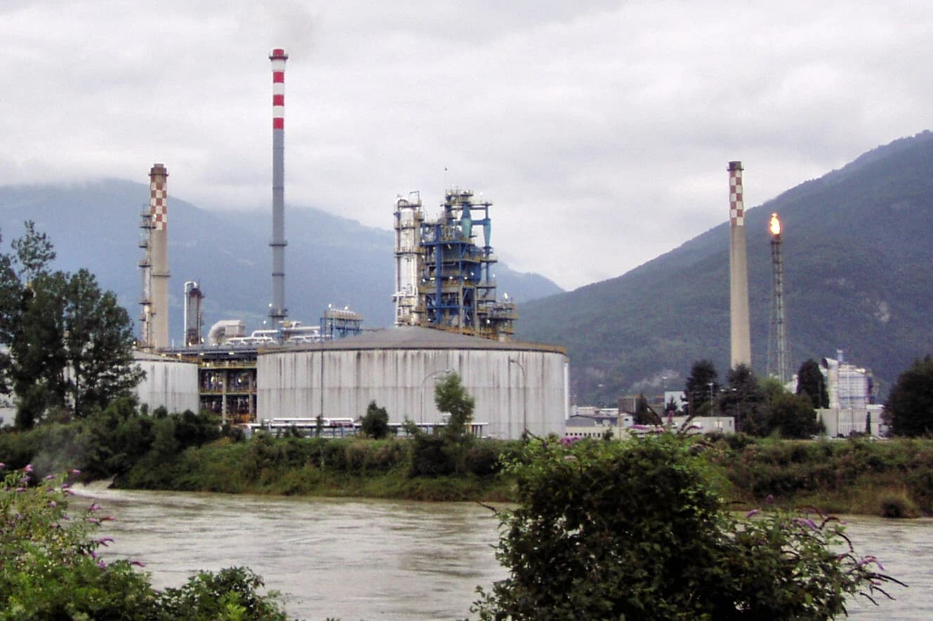 Industrial refinery with multiple towers, storage tanks, and a flaring chimney next to a river.
