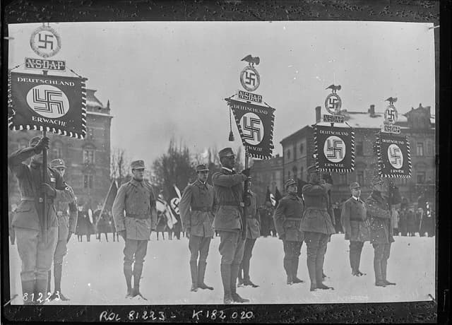 Men in military uniforms holding flags with Nazi symbols during a ceremony in Munich.