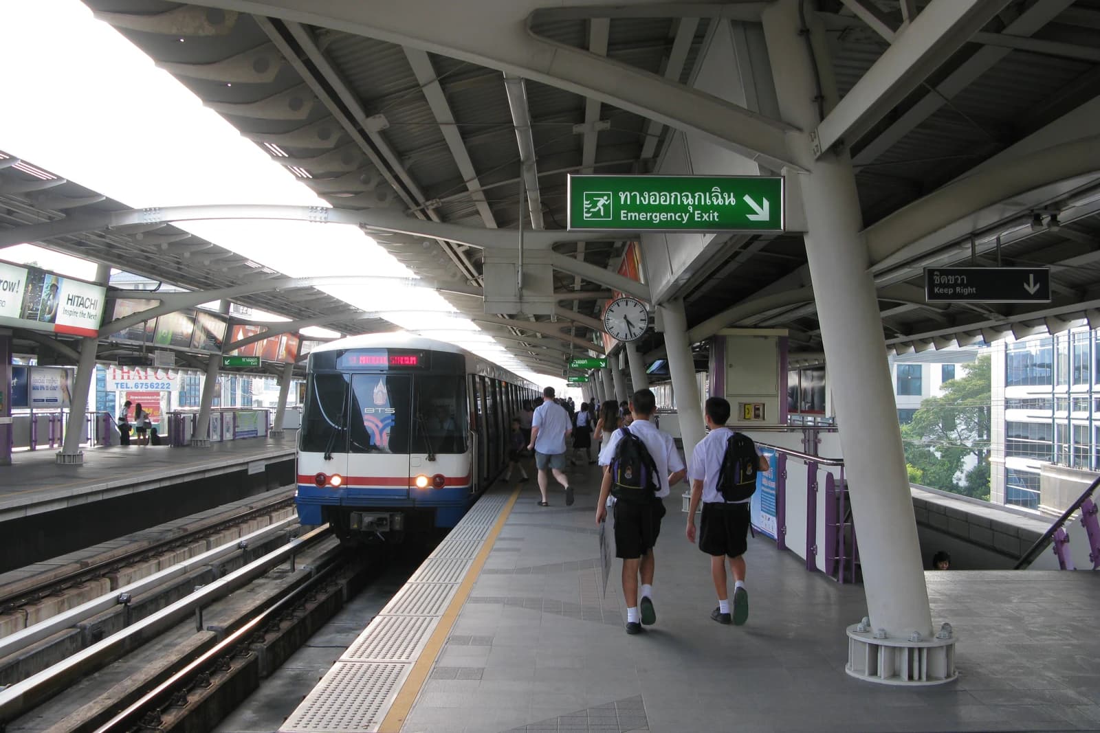 BTS Skytrain approaching a station platform with passengers walking in Bangkok, Thailand.