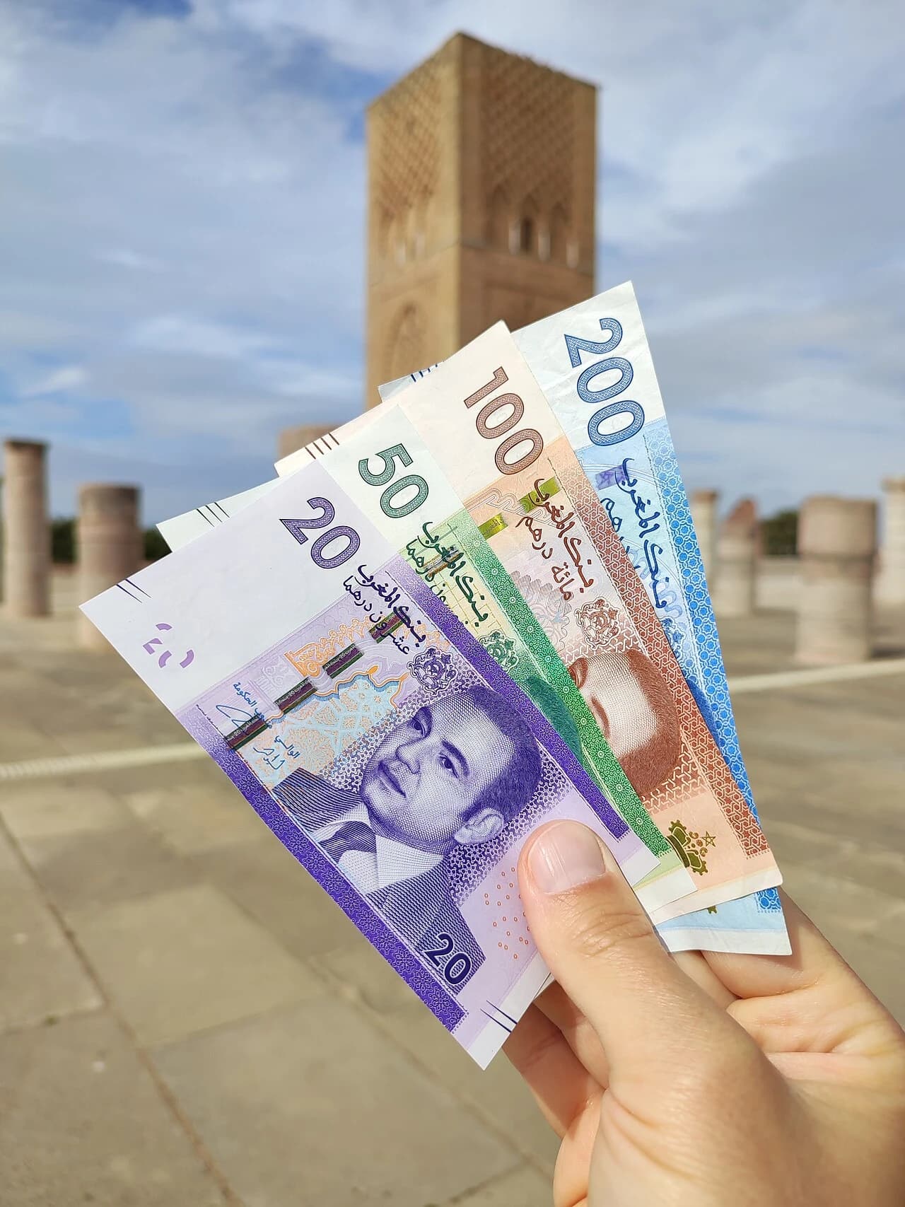 Hand holding various denominations of Moroccan dirham banknotes in front of Hassan Tower.