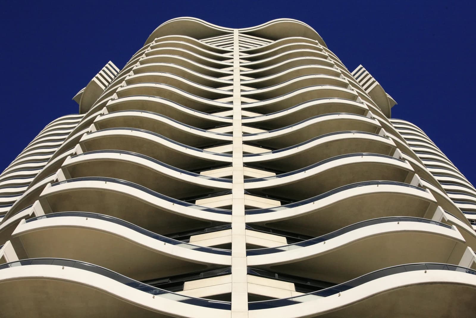 Modern high-rise apartment building with wavy balconies viewed from below against a deep blue sky.