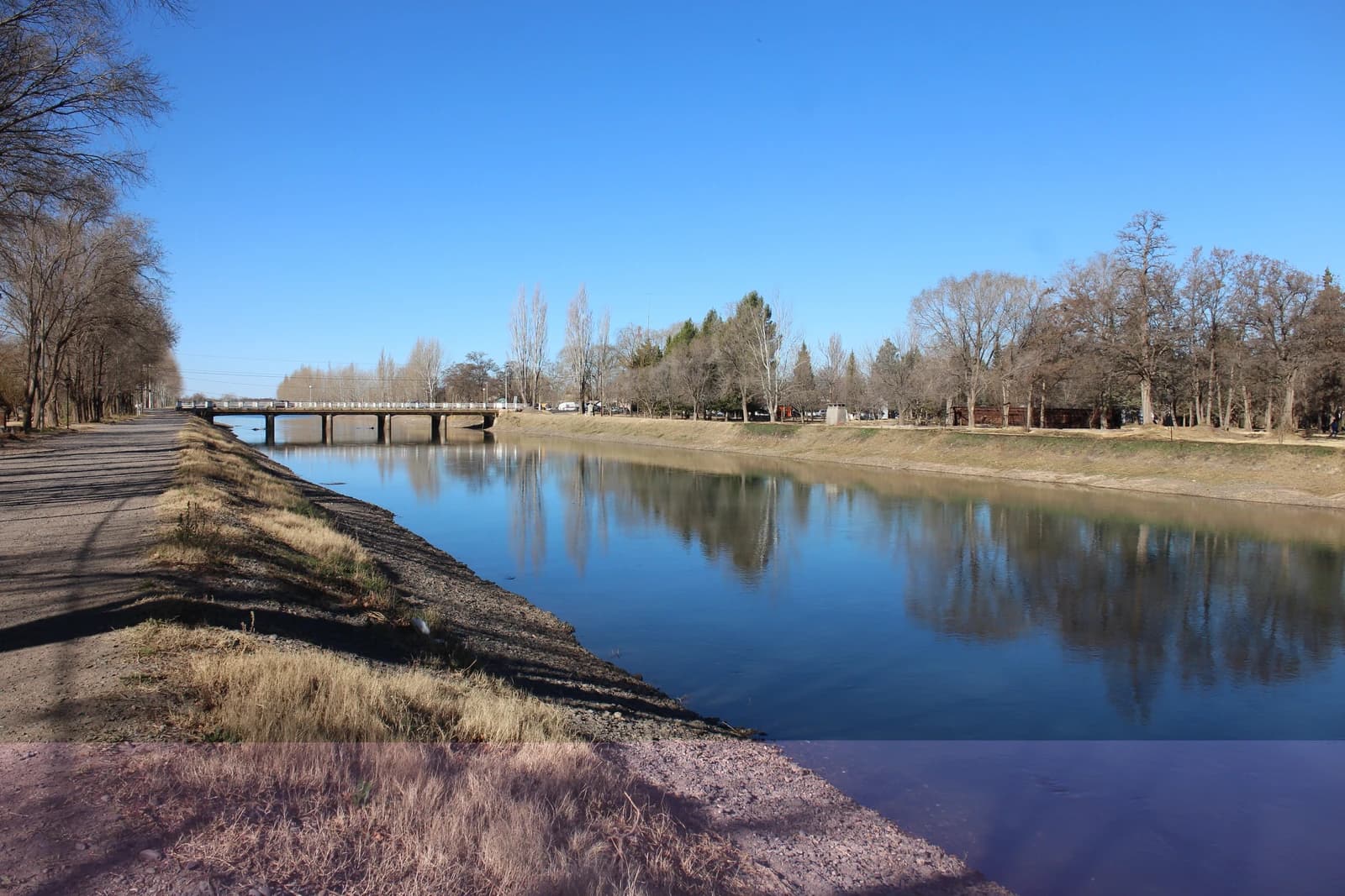 A road bridge crosses a wide irrigation canal with trees and a dirt path alongside it.