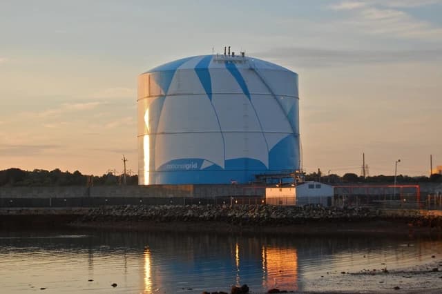 A large National Grid LNG storage tank, painted white and blue, seen at sunset with reflections in the water.