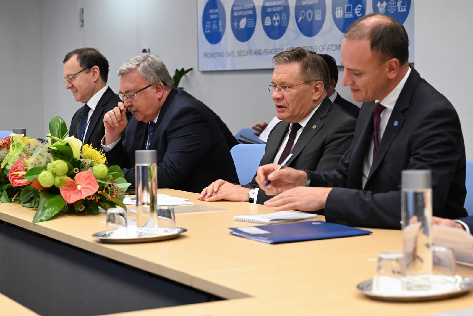 Four men in suits sitting at a conference table, with one speaking.
