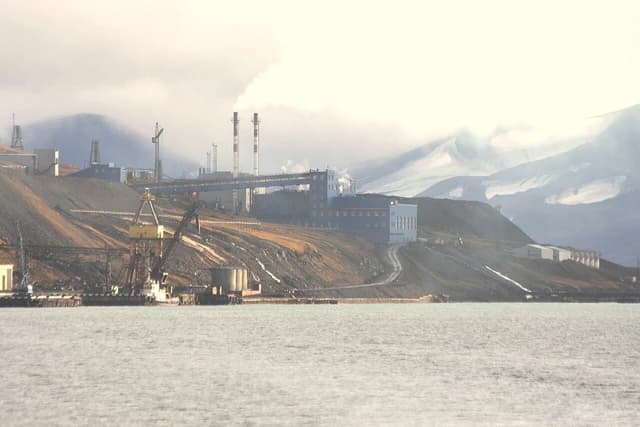 Coal power plant with smoking chimneys and mining operations alongside a body of water in Barentsburg.