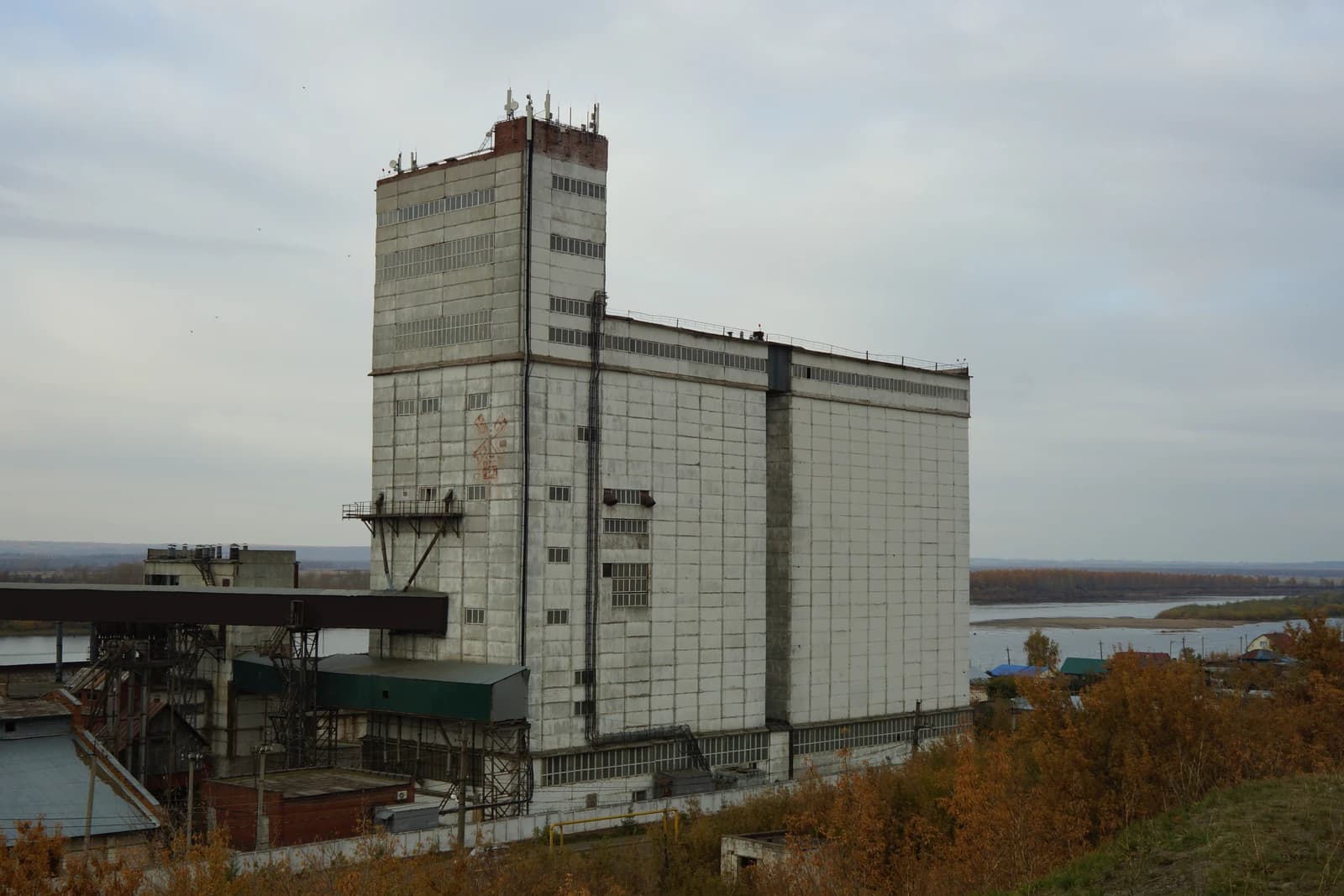 Large concrete grain elevator building with a river and autumn trees in the background.