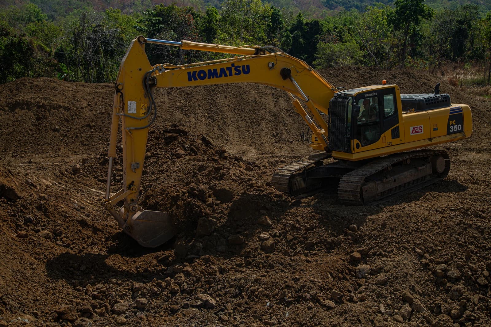 Komatsu excavator digging earth on a brown dirt hill, with green trees in the background.