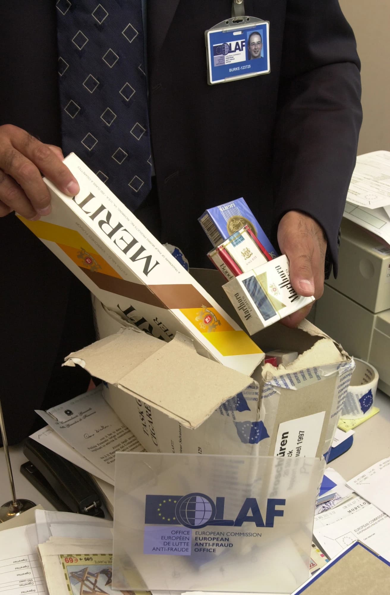 An OLAF official's hands inspecting boxes of cigarettes at a desk.