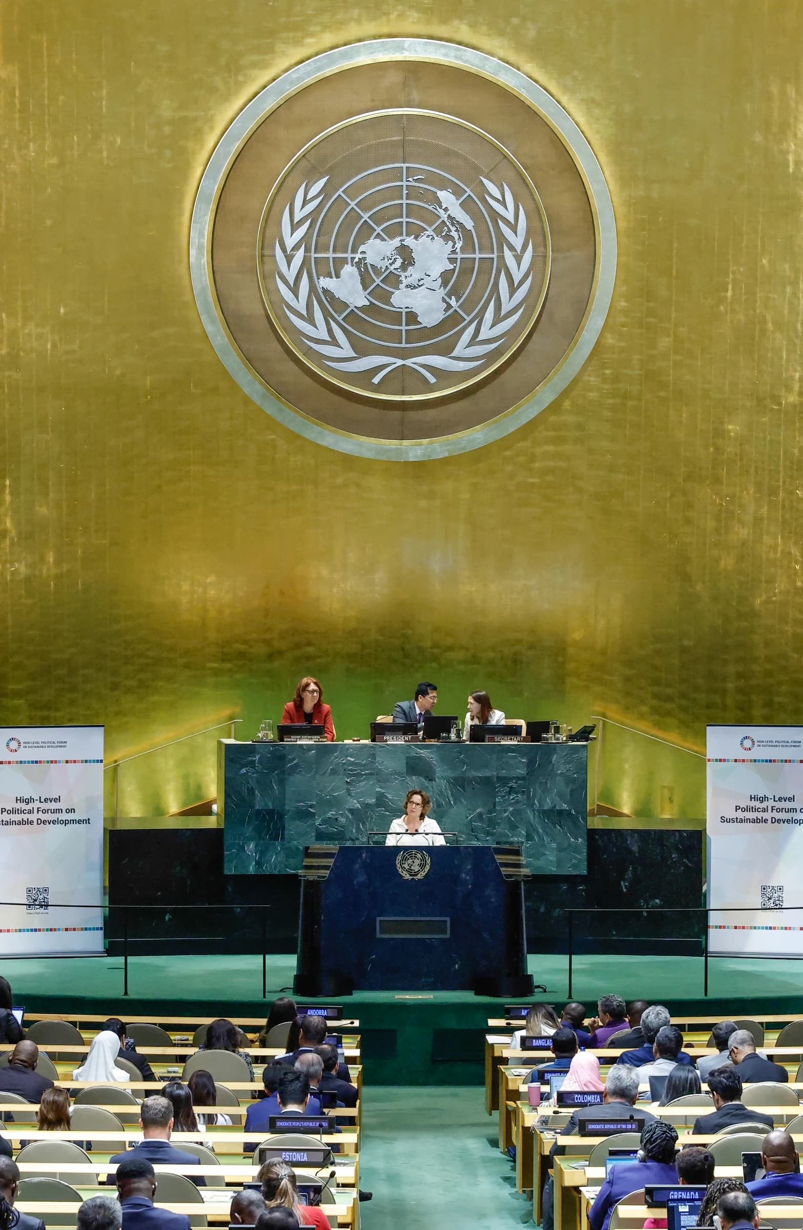 A woman speaks from a podium at the front of a UN General Assembly hall.