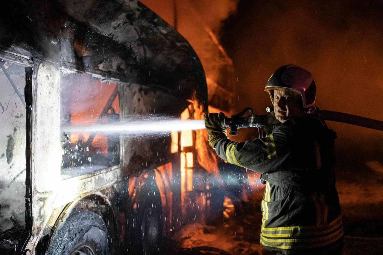 Firefighter wearing helmet and protective gear, spraying water from a hose at a burning vehicle.