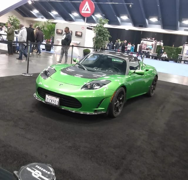 Bright green Tesla Roadster with a dark grey hood on display indoors at an auto show.