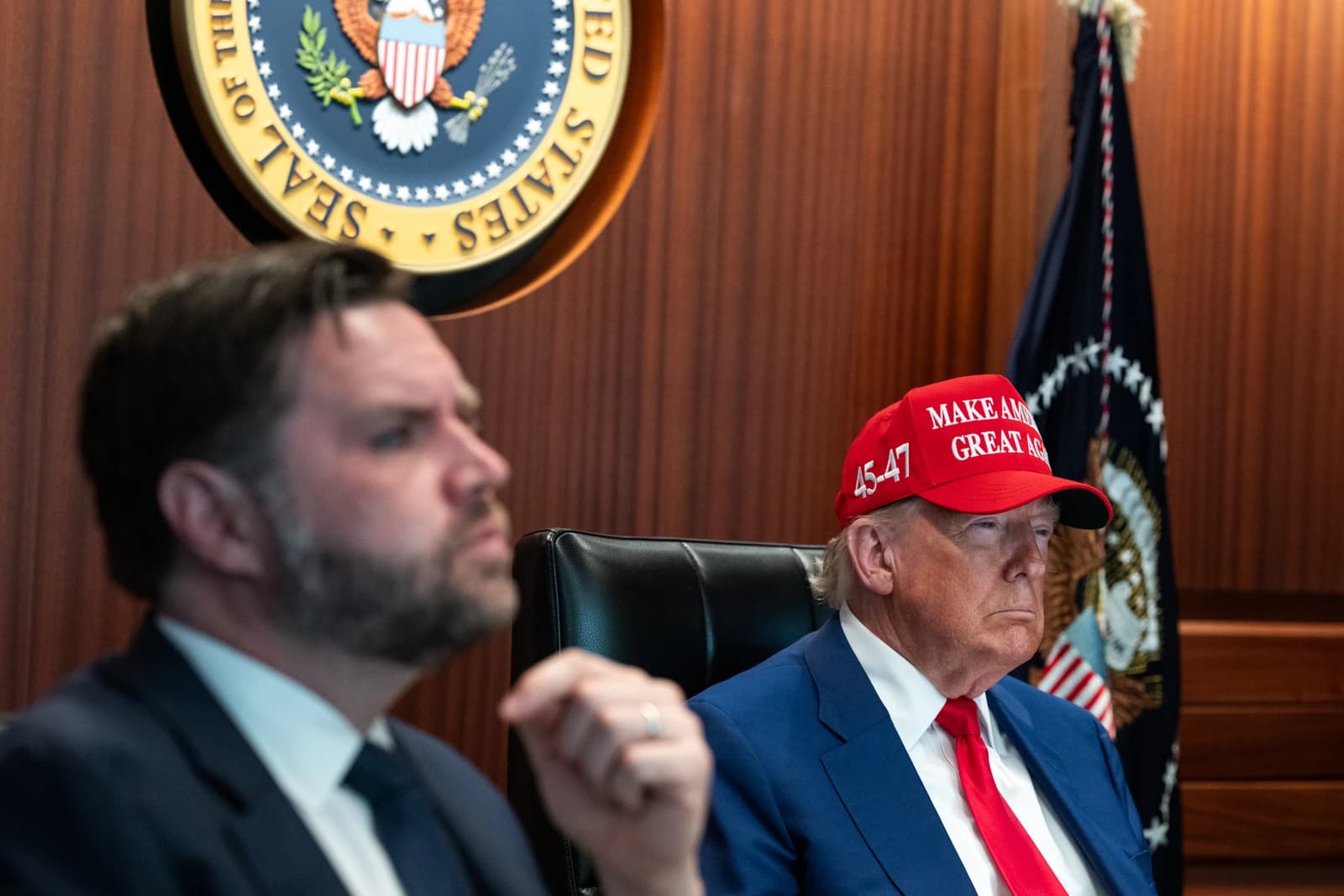Former President Donald Trump seated during a meeting, wearing a red hat.
