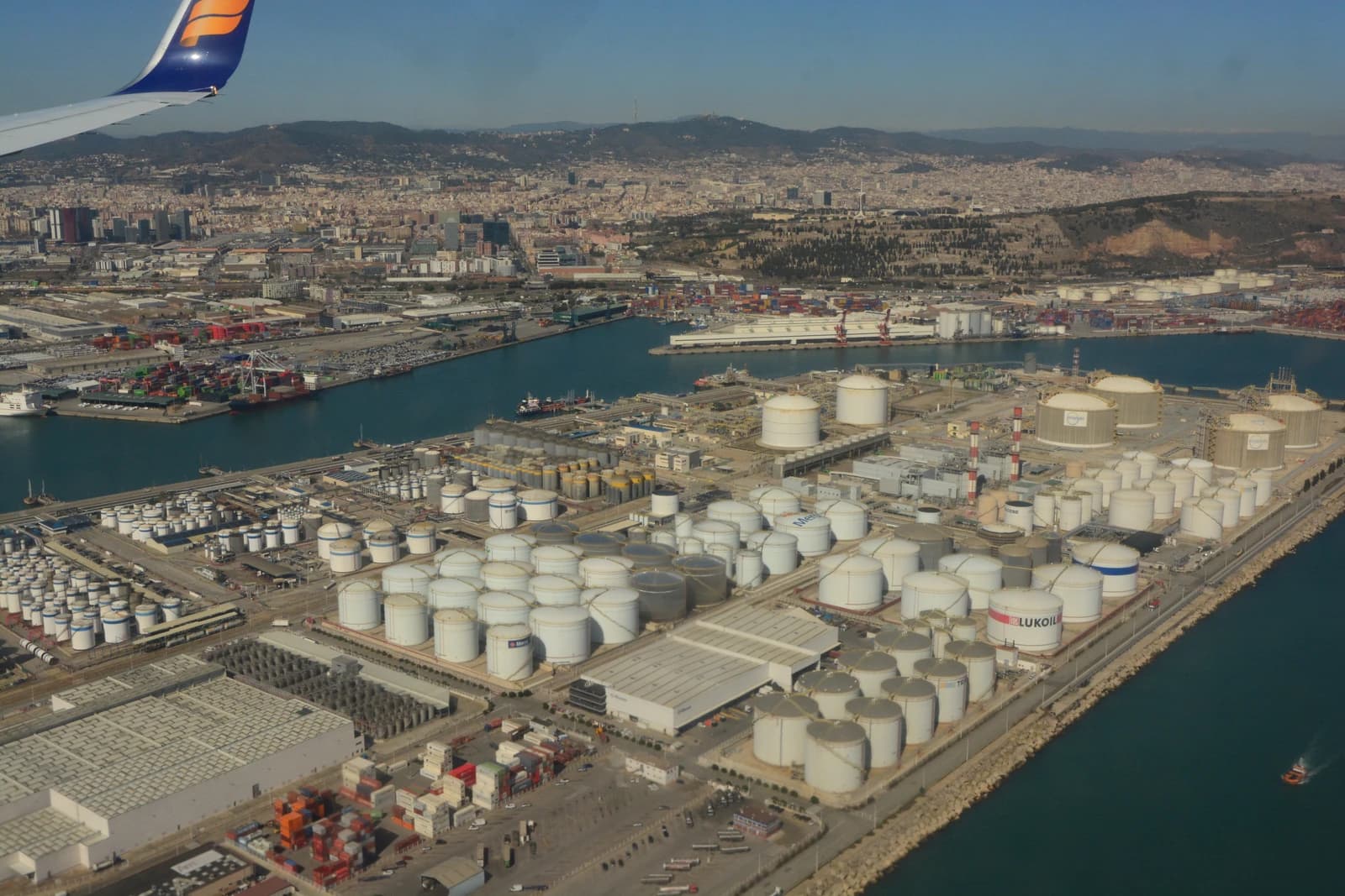 Aerial view showing numerous white oil storage tanks and industrial facilities next to a body of water in Barcelona.