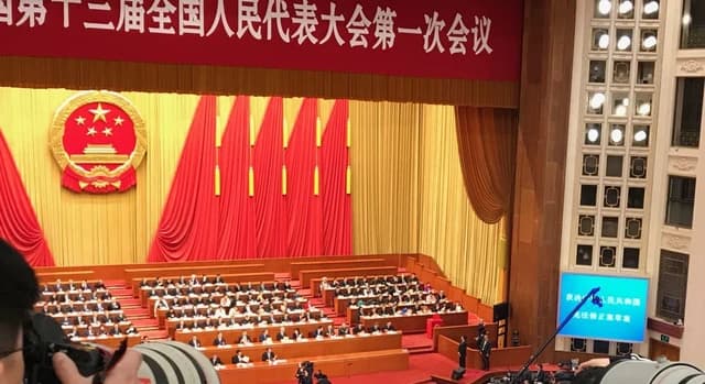 Delegates seated in a large hall under the Chinese national emblem at a formal session.