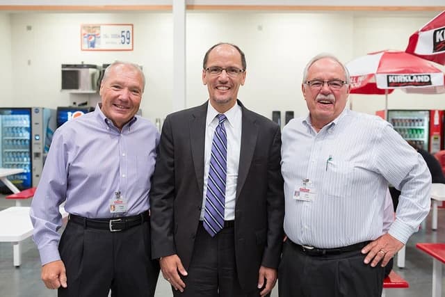 Three men, including one in a suit and tie, smiling and standing indoors.