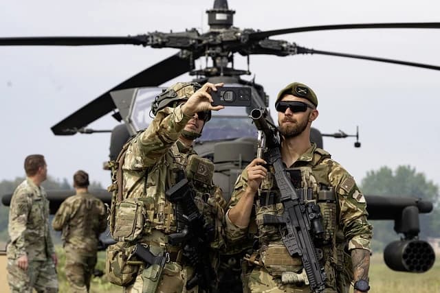 Two soldiers, one taking a photo, with an Apache helicopter in the background.