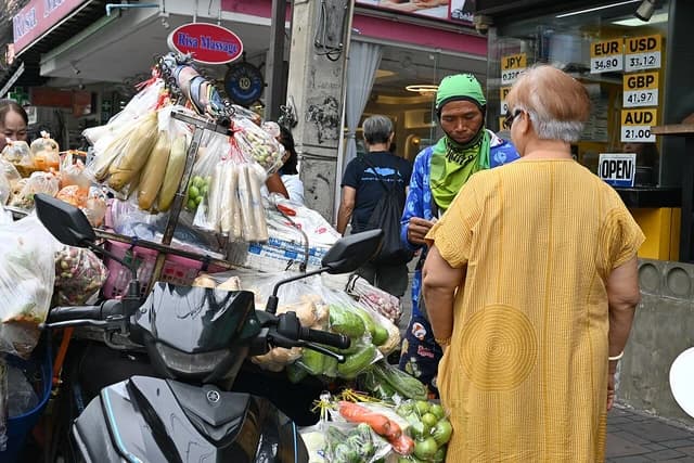 A street vendor with produce strapped to a motorbike talks to a customer near a currency exchange sign.