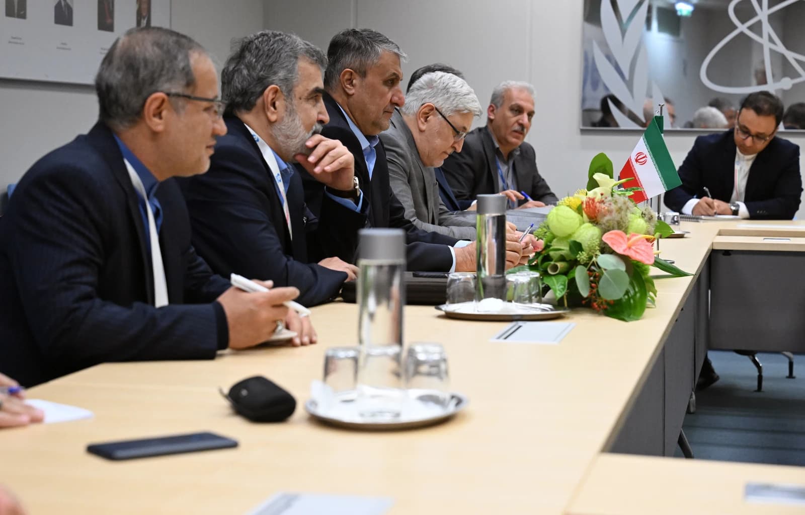 Iranian officials seated at a conference table with an Iranian flag, taking notes.