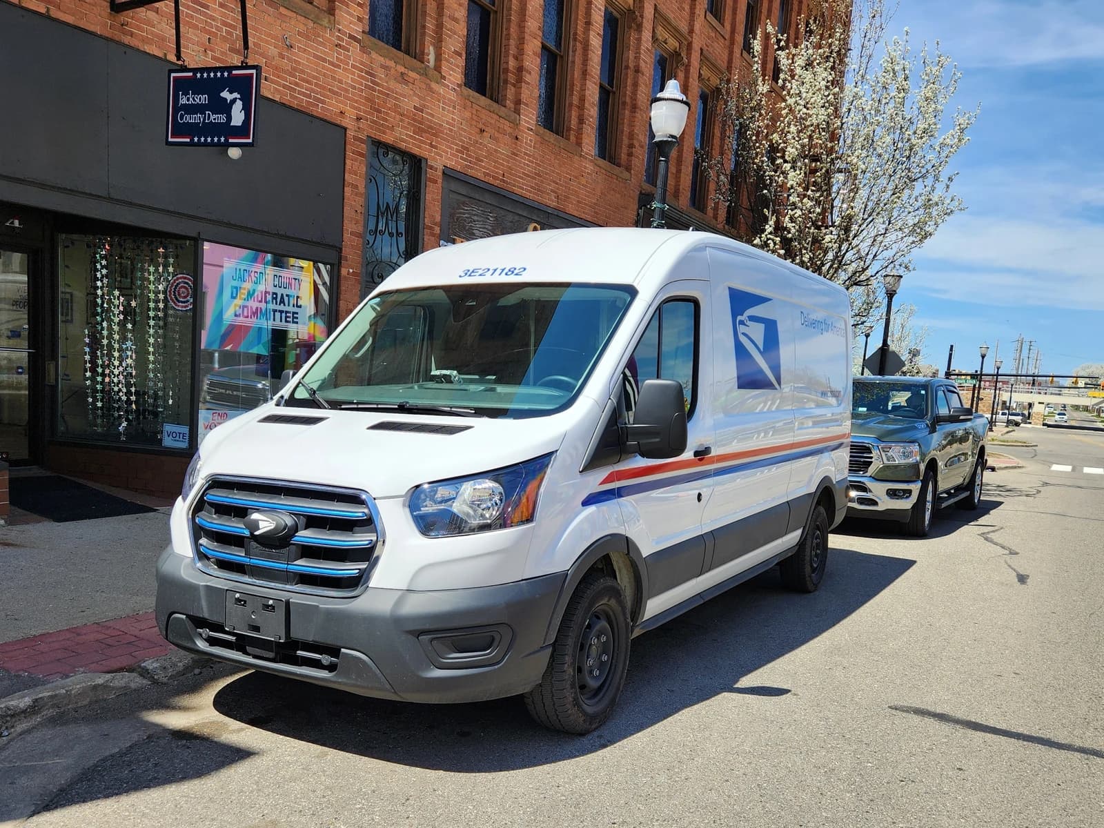 A white USPS Ford E-Transit electric van parked on a city street.