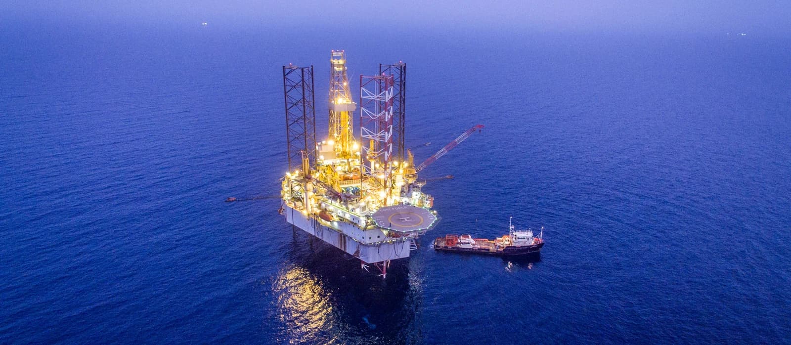 Aerial view of a brightly lit offshore gas drilling platform in the ocean at dusk.