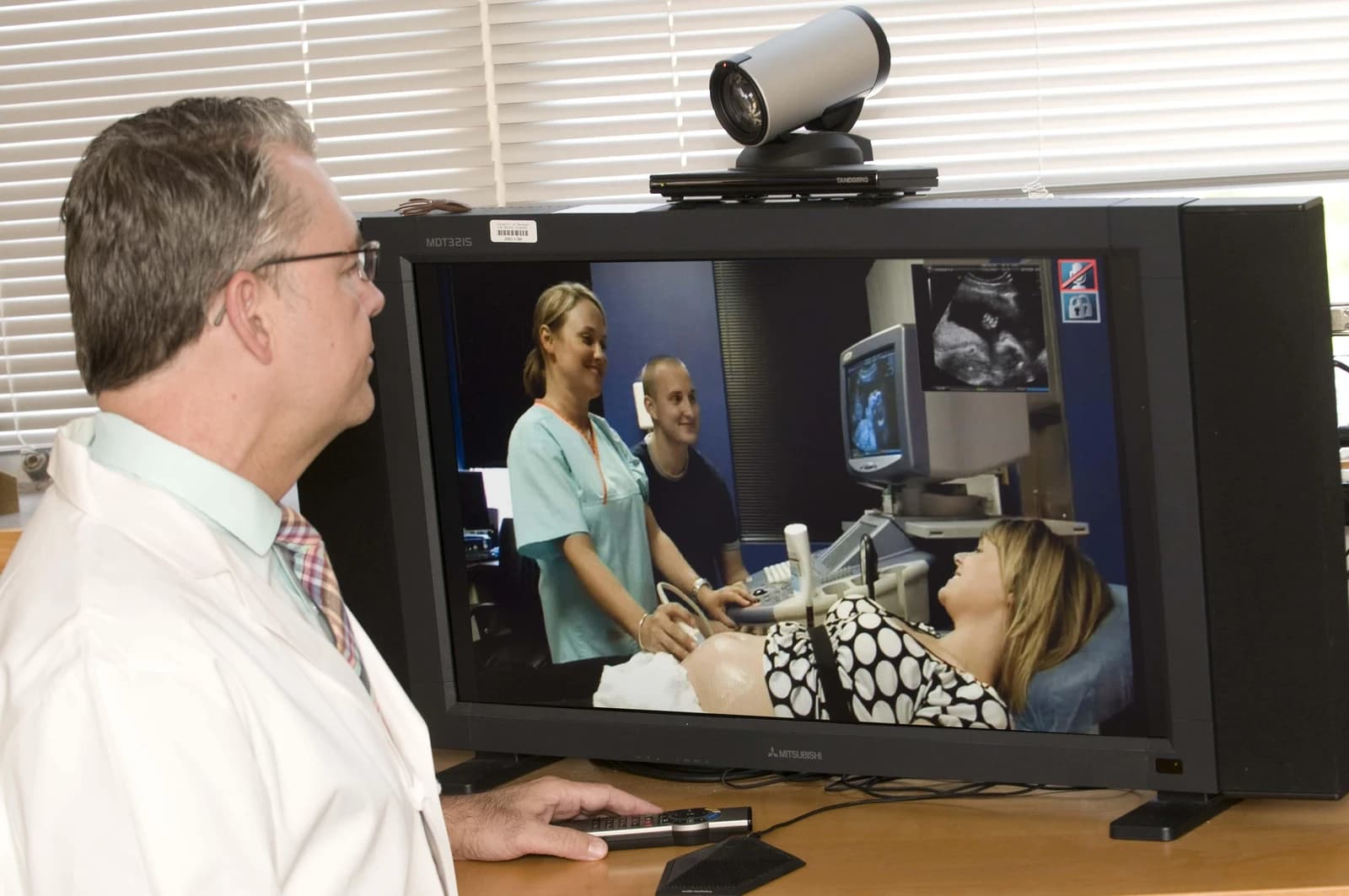 Man in white lab coat watching a video conference of a medical examination on a large monitor.