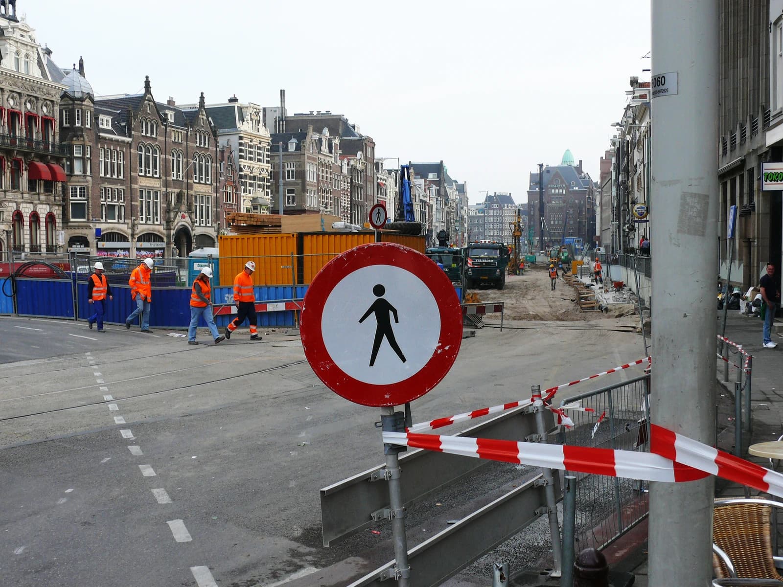 Construction workers in orange vests and hard hats walking on a building site in Amsterdam.