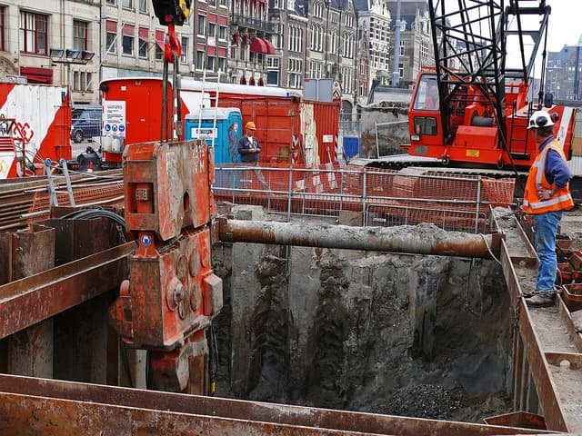 Construction workers in hard hats and safety vests supervising sheet piling activities in a trench.