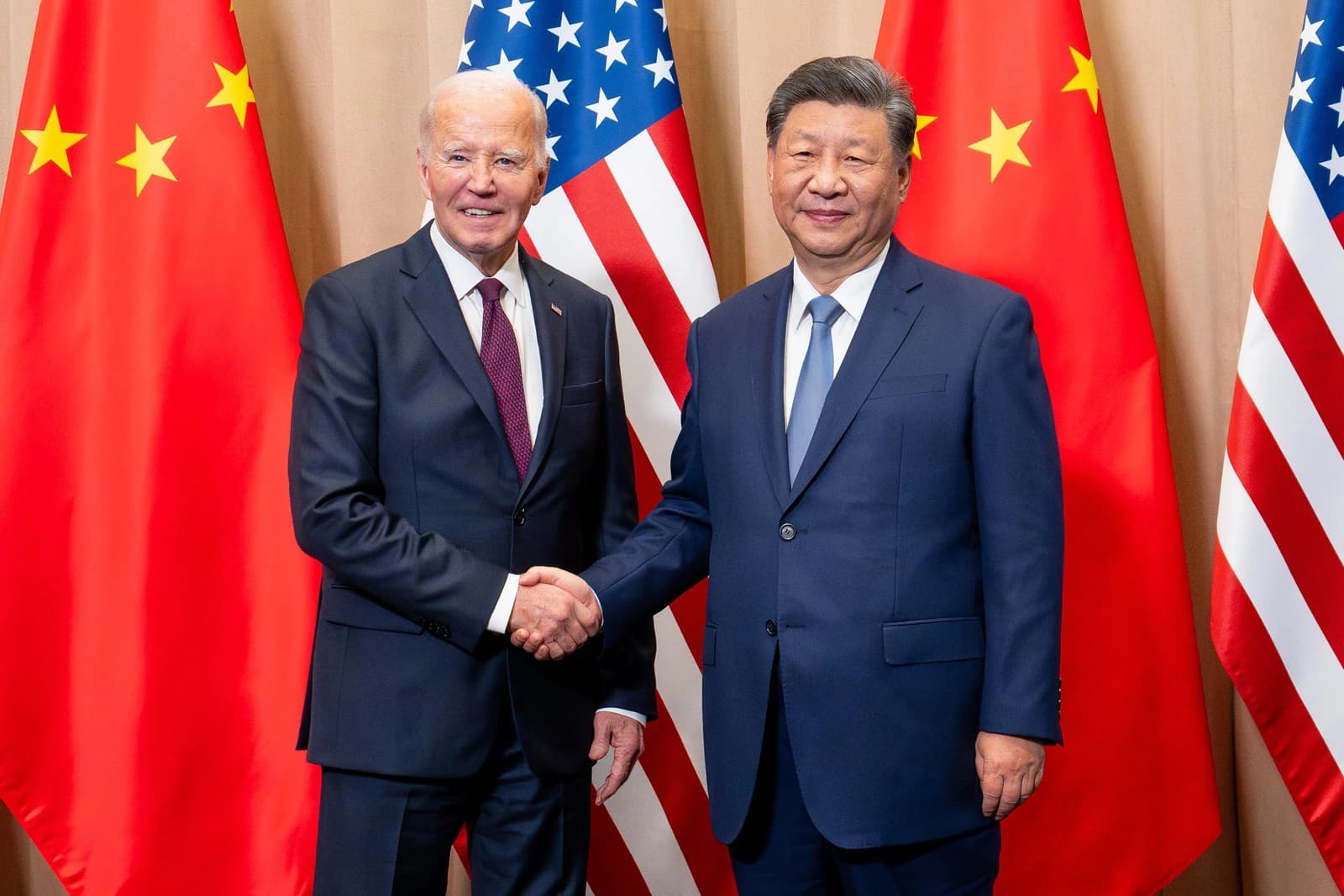 President Joe Biden and President Xi Jinping shake hands in front of US and Chinese flags.
