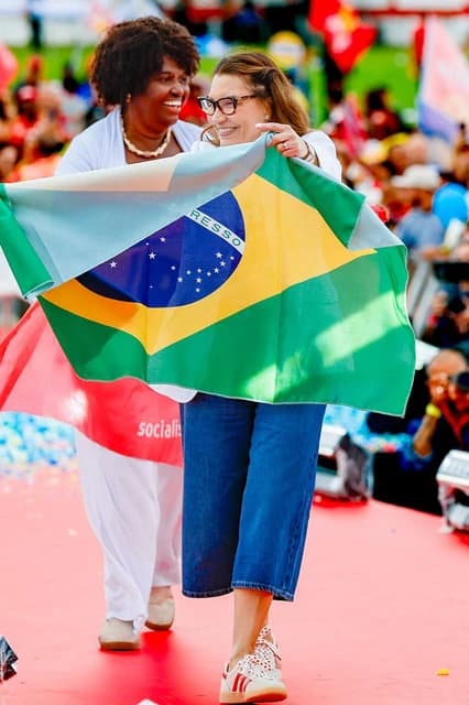 Two women, one in glasses, holding a large Brazilian flag at an event.