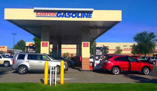 Several vehicles are lined up at a Costco gas station with the store visible in the background.