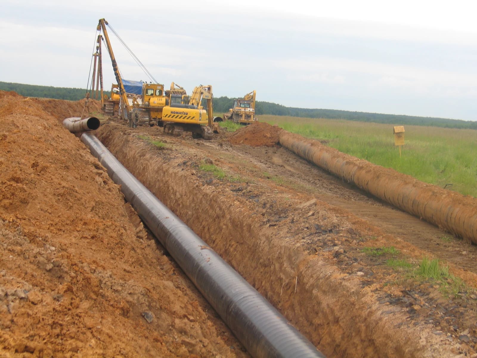 Construction machinery installing large diameter pipelines in an open trench in a field.