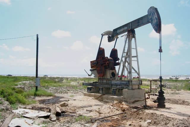 An oil pumpjack stands prominently near a sandy beach under a blue sky.