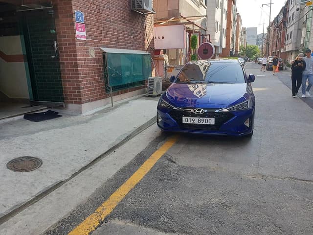 A blue Hyundai Elantra parked on a narrow street with buildings and pedestrians in the background.