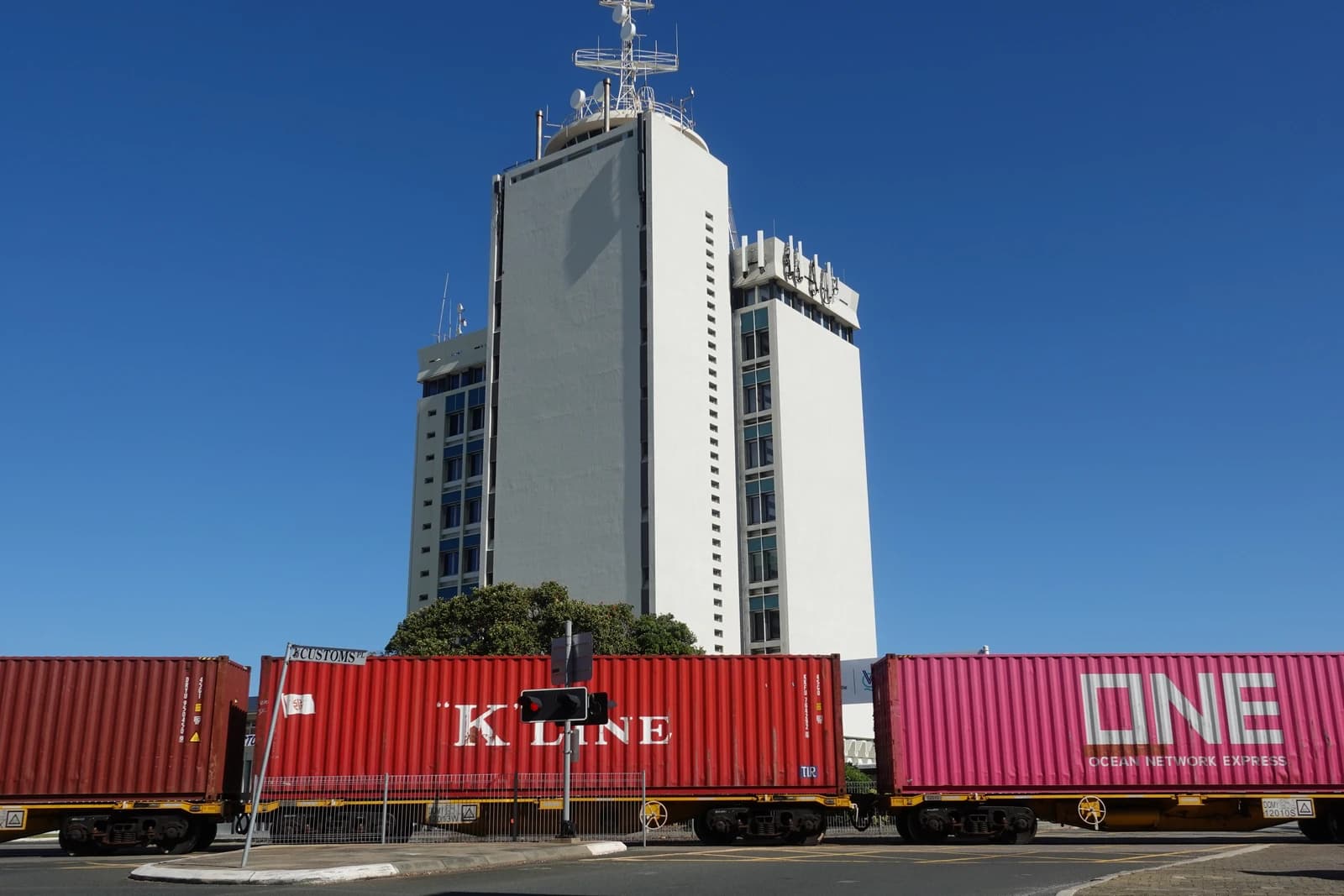 Train carrying red and pink shipping containers passing a tall white building.