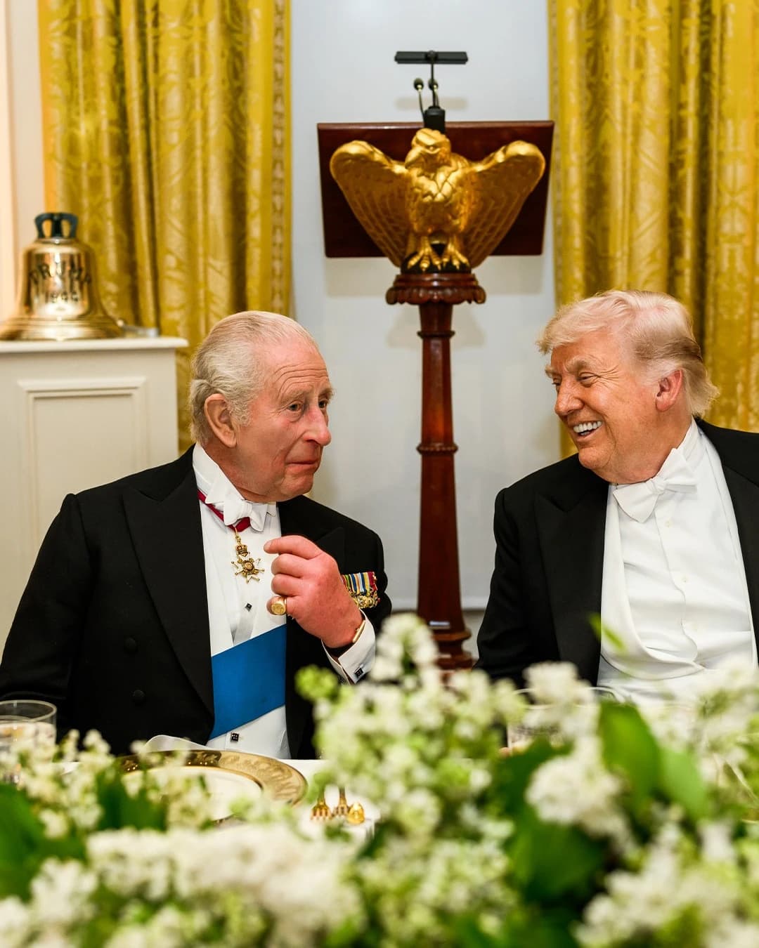 King Charles III and Donald Trump at a formal dinner setting, looking towards each other.
