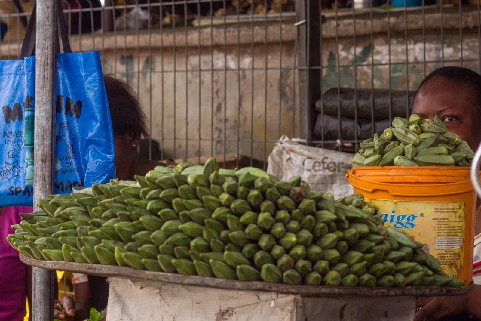 Okra arranged for sale on a tray at a market stall, a person partially visible in background.